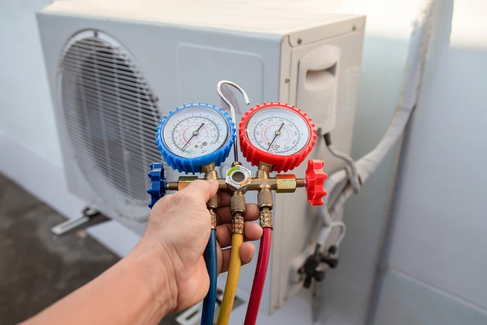 Technicien tenant un manifold de climatisation avec deux manomètres à cadran bleu et rouge, connecté par des flexibles colorés devant une unité extérieure de climatisation résidentielle