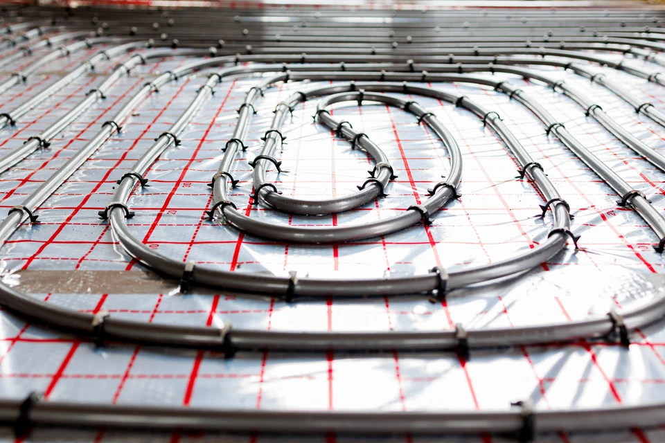 Installateur en combinaison beige travaille sur système de plancher chauffant hydraulique, connectant tuyaux PEX au collecteur-répartiteur avec vannes rouges et bleues